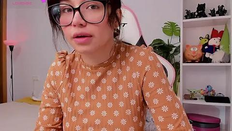 Media: Video of a young woman with fair skin, wearing black-rimmed glasses and a floral-patterned brown top, seated on a pink gaming chair in a cozy, colorful room.