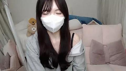 Media: Video of a young woman with long dark hair, wearing a white face mask, a gray sweater, and a white off-shoulder top, sitting on a plush pink sofa in a cozy room with stuffed toys and a mirror.