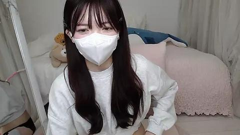 Media: Video of a young Asian woman with long, straight black hair, wearing a white mask, white long-sleeved shirt, and sitting on a bed with stuffed animals and pink blankets.