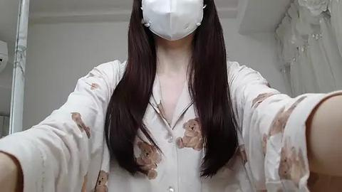 Media: A video of a woman with long, straight dark hair, wearing a white shirt with teddy bear pattern, a white face mask, and white gloves, in a sterile room with white walls and a white gown hanging.