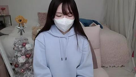 Media: A video of an Asian woman with long black hair, wearing a white face mask, a light blue hoodie, and a cozy living room with pink pillows, a Christmas tree, and a teddy bear.