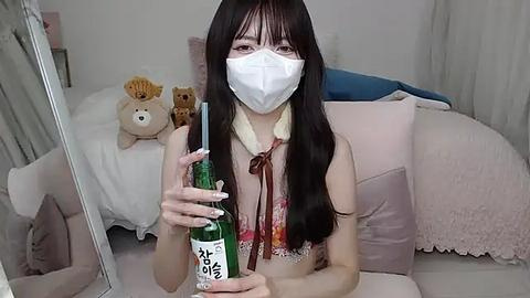 Media: Video of a young East Asian woman with long black hair, wearing a white mask and a colorful floral blouse, holding a green bottle, in a cozy, softly lit room with plush toys and pillows.