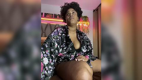 Media: Video of a plus-sized Black woman with natural hair, wearing a floral kimono, sitting on a bed. Background features a cozy, dimly lit room with warm lighting and potted plants.