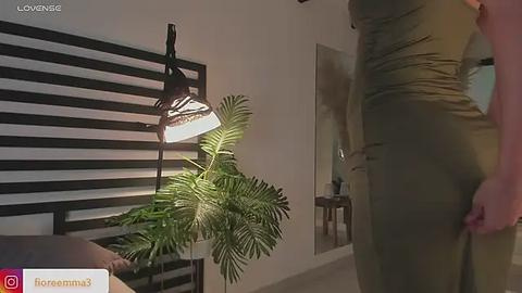 Media: Video of a woman in a tight, olive-green dress adjusting her waistband, standing in a modern room with a black and white striped wall, a large potted fern, and a warm-toned light fixture.