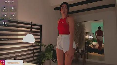 Media: Video of a slim, dark-haired woman in a red crop top and white shorts, standing in a modern bedroom with a large mirror reflecting her image.