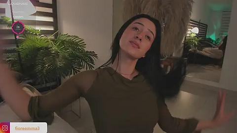 Media: A video of a young woman with long black hair, wearing a sheer green top, taking a selfie in a modern, cozy living room with greenery and a plush rug.