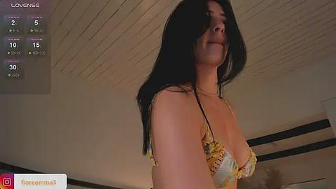 Media: Video of a light-skinned woman with long black hair, wearing a yellow floral bra, captured from a low angle in a dimly lit room.