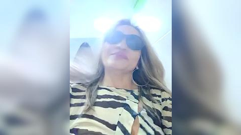 Media: A video of a woman with long blonde hair, wearing large black sunglasses, a leopard-print blouse, and a silver necklace, smiling. The background is blurry, with a bright, light blue ceiling.