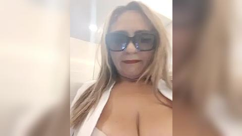 Media: Video of a blonde, middle-aged woman with large glasses and a white shirt, showing cleavage, taken from a low angle, slightly blurred.