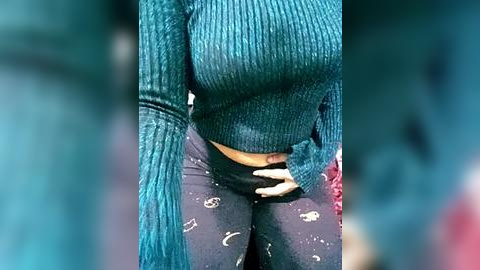 Media: Video of a woman in a teal, ribbed, long-sleeved sweater and dark leggings adorned with white celestial patterns. Her hands rest on her waist, showcasing her midsection.