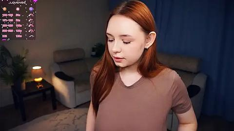 Media: Video of a young, fair-skinned woman with straight, reddish-brown hair, wearing a light brown T-shirt, standing indoors with a dimly lit room in the background.