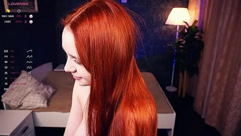 Media: Video of a fair-skinned woman with long, straight red hair, seen from the side in a dimly lit bedroom. She is topless, with a soft smile. A lamp and a bed with a white duvet are in the background.