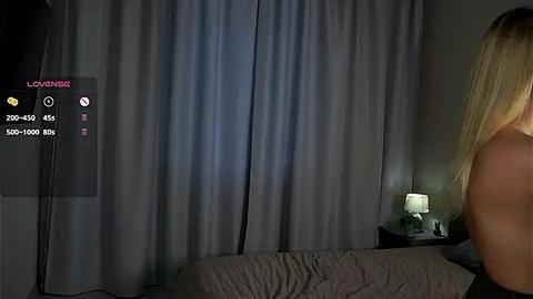 Media: A dimly lit bedroom scene with a blonde woman's back, a bed with white sheets, a nightstand lamp, and gray curtains.