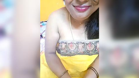Media: Video of a smiling, dark-skinned woman with medium-length black hair, wearing a yellow strapless top with intricate embroidery and a gold necklace.