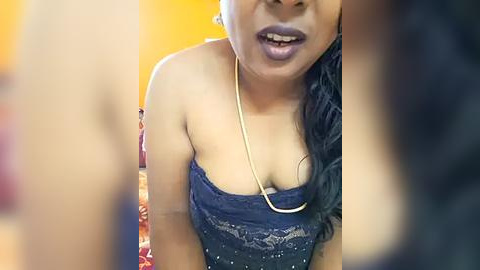Media: A close-up video of a South Asian woman with medium brown skin and long black hair, wearing a strapless blue lace dress. She has a gold necklace and is smiling, with blurred figures in the background.