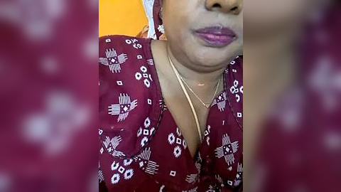 Media: Video of a middle-aged Black woman with medium-dark skin, wearing a burgundy blouse with white geometric patterns, and gold necklace. She has a neutral expression, with a hint of a smile. The background is a blurred yellow-orange color.