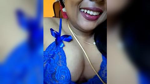 Media: A close-up video of a woman with medium brown skin, wearing a revealing blue lace bra with a gold necklace, smiling broadly. Background is blurred.