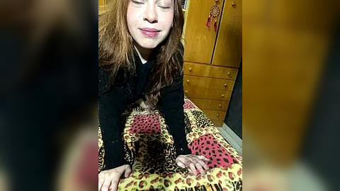 Media: Video of a young woman with long, wavy red hair, wearing a black sweater, lying on a leopard-print blanket, in a cozy, warmly lit room with wooden cabinets and a red tassel in the background.