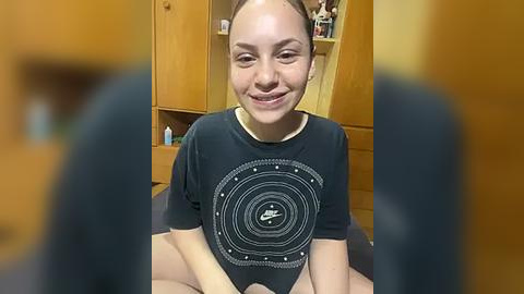 Media: Video of a smiling Caucasian girl with light skin, brown hair, and a Nike T-shirt in a bedroom with wooden furniture and a blurred view of a person's legs.