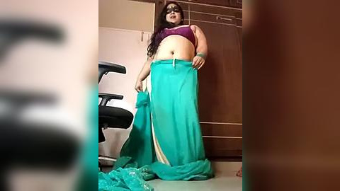 Media: Video of a plus-size woman in a teal mermaid costume, holding a matching tail, standing in a room with a black chair and brown wooden cabinets.