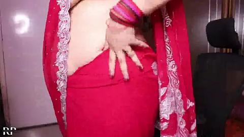 Media: A video shows a fair-skinned woman in a red saree, with intricate white floral embroidery, adjusting her blouse. Her right hand, adorned with colorful bangles, pulls the blouse aside. The background features a black sofa and a white door.