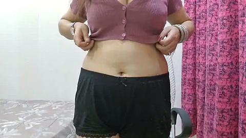 Media: Video of a woman lifting a pink, ribbed crop top to reveal a midriff, wearing black pajama shorts and multiple bracelets. Background features a pink, patterned curtain and a tiled floor.