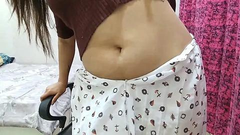 Media: Video of a woman with light brown skin and long dark hair, lifting her maroon crop top to reveal her flat stomach, wearing white pants adorned with small, colorful designs. Background includes a bed and a pink, patterned curtain.