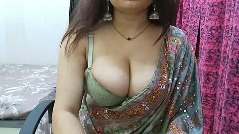 Media: A video of an Asian woman with fair skin, wearing a green blouse with a plunging neckline, revealing cleavage. She has long, dark hair and is adorned with dangling earrings. Background includes a bed with floral bedding and a pink, textured curtain.