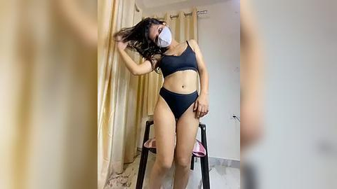 Media: Video of a young Asian woman in a black sports bra and high-waisted shorts, wearing a white face mask. She stands in a bedroom with beige curtains and a white door, holding her long black hair.