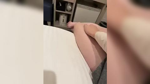 Media: Video of a person's legs and lower torso, lying on a white bed with beige socks, in a dimly lit room with a white cabinet and laundry basket in the background.