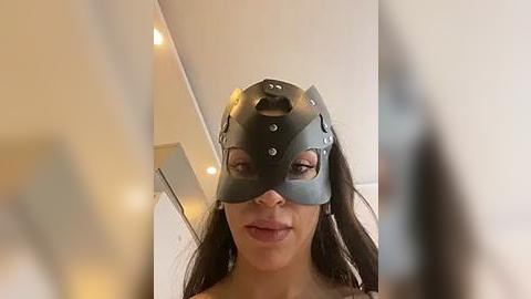 Media: Video of a woman wearing a black, leather BDSM mask with studs, smiling. Background includes a white ceiling and blurred, dimly lit objects.