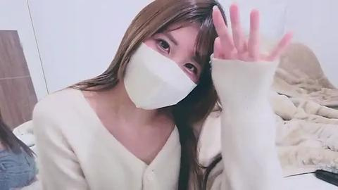 Media: Video of a young Asian woman with long brown hair, wearing a white face mask, beige cardigan, and a white long-sleeve shirt, waving her hand in a casual bedroom setting with beige sheets.