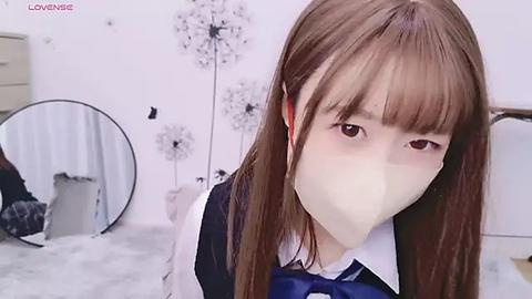 Media: Video of an Asian girl with long brown hair, wearing a white shirt and blue vest, masked, standing in a white room with floral decor, round mirror, and furniture.