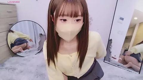 Media: Video of an Asian woman with long straight brown hair and a yellow mask, wearing a yellow cardigan, kneeling on a gray carpet in a bedroom with a round mirror and wooden furniture.