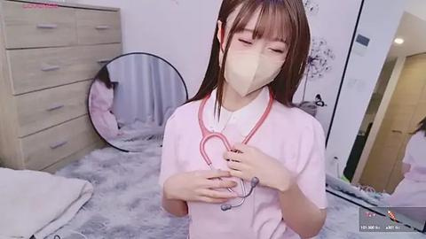 Media: Video of an Asian woman with long brown hair, wearing a pink shirt, white mask, and red stethoscope, in a cozy bedroom with a fluffy rug, wooden dresser, and round mirror.