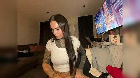 Media: A video of a tattooed woman with long black hair, wearing a white t-shirt, sitting on a brown couch in a dimly lit living room, watching TV.