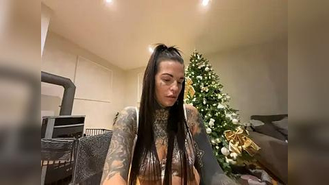 Media: Video of a tattooed woman in a sleeveless top, seated indoors near a decorated Christmas tree; blurred background.