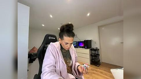 Media: Video of a woman with dark hair in a bun, wearing a lavender hoodie, working at a black gaming chair in a minimalist, brightly-lit room with a wooden floor and white walls.