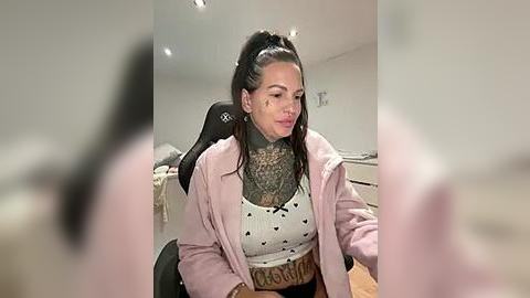 Media: Video of a tattooed woman with a high ponytail, wearing a pink blazer over a white tank top, sitting at a desk in a modern room with white walls and wooden floors.