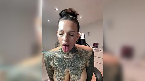 Media: A video of a tattooed woman with dark hair in a bun, wearing a white dress, sticking out her tongue, taken in a modern room with white walls and a desk.