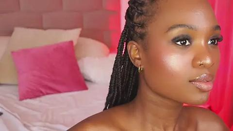 Media: Video of a young Black woman with smooth, dark skin, wearing gold hoop earrings, braided hair, and a subtle makeup look. She is in a bedroom with pink and white decor.