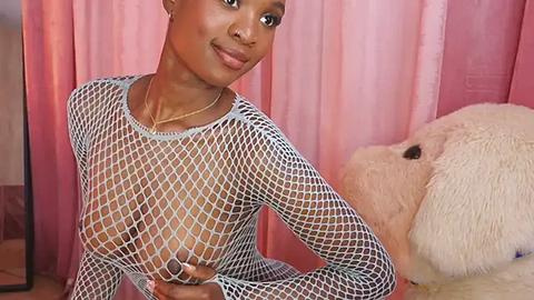 Media: Video of a slender, dark-skinned woman with short hair, wearing a fishnet top that reveals her small breasts, standing beside a large, plush teddy bear in a pink-tiled room.