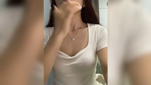 Media: Video of a woman with long brown hair, wearing a white T-shirt, with a hand covering her mouth, blurred background.