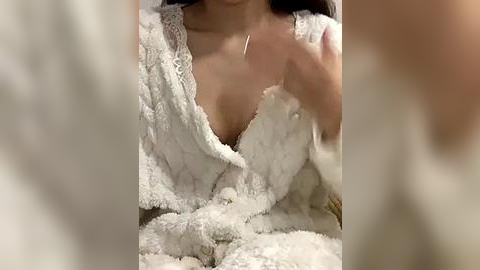 Media: Video of a woman in a white, fluffy bathrobe, holding the robe open to reveal her cleavage and a thin necklace. The background is blurred, focusing on her partially visible face.