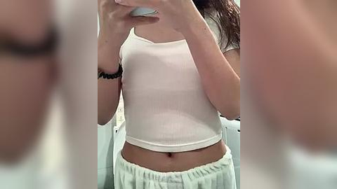 Media: Video of a young woman with light skin, wearing a white cropped tank top and white pajama pants, taking a selfie in a bathroom, blurred background.