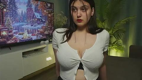 Media: Video of a young woman with long dark hair, wearing a white ribbed crop top, sitting in a cozy living room with a Christmas-themed TV screen and greenery background.