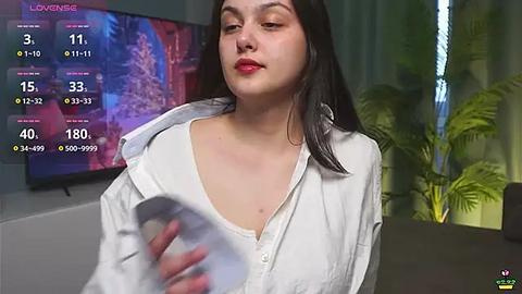 Media: Video of a young woman with fair skin, red lipstick, and long black hair, wearing a white shirt, holding a white object. Background features a TV displaying a colorful image and a green potted plant.