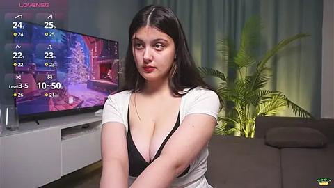 Media: Video of a young, light-skinned woman with long black hair, wearing a low-cut white top, sitting on a couch. Background features a TV screen displaying a virtual reality game, a green plant, and a dark sofa.