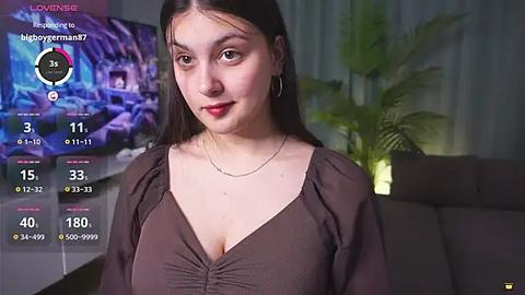 Media: Video of a fair-skinned, young woman with long brown hair, wearing a low-cut, brown top, standing indoors with a TV showing a game in the background.