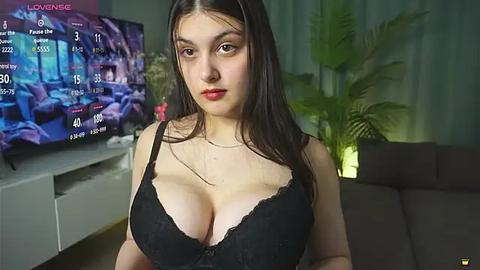 Media: Video of a young woman with long dark hair, fair skin, and large breasts in a black lace bra, standing in a living room with a TV displaying a football game and green curtains.